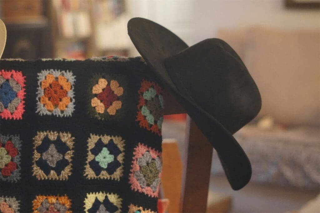 Black afghan with colorful squares draped over the back of a wooden rocking chair with a black cowboy hat propped on the corner
