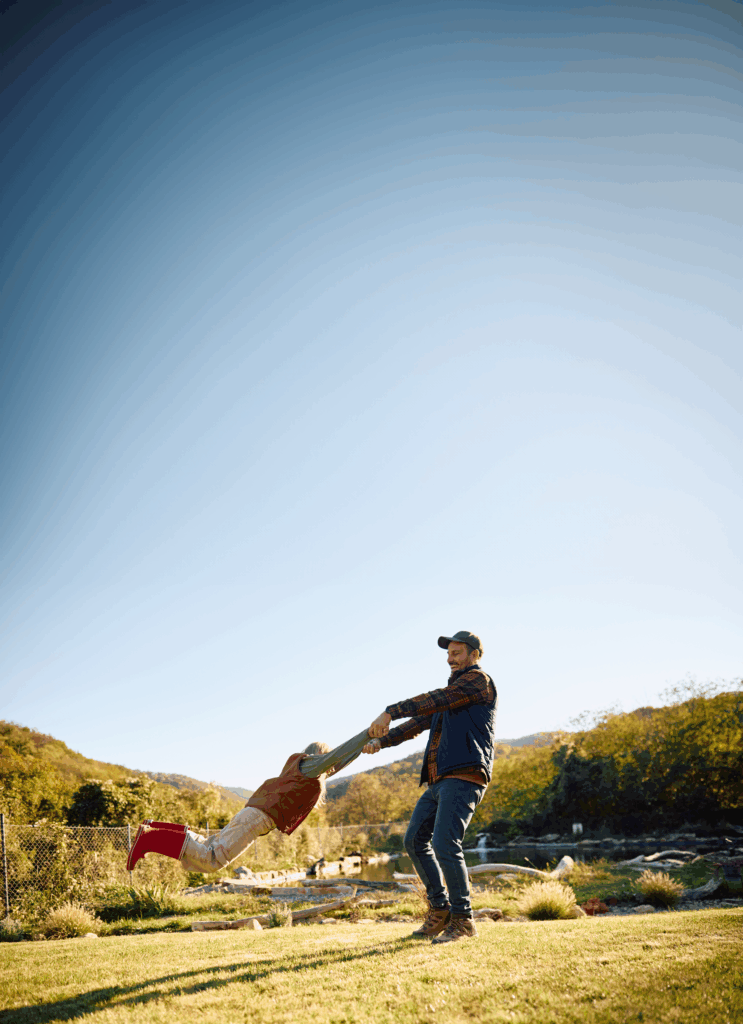 Happy father in a plaid shirt and vest and jeans spinning his young daughter, wearing red boots and a tan vest around in a green field with a bright blue sky
